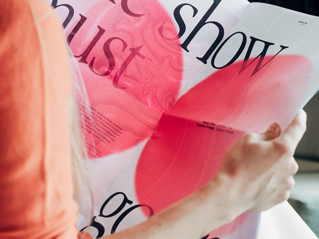 a-blonde-woman-photographed-over-the-shoulder-holds-a-newspaper-sized-magazine-with-neon-pink-details