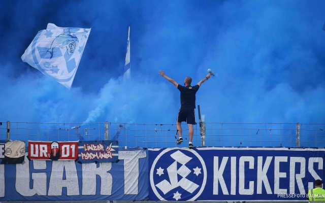 a-man-stands-on-a-wall-which-is-covered-with-stuttgarter-kickers-banners-the-fan-block-is-full-of-blue-smoke