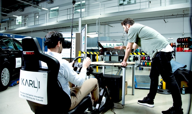 zwei-Leute-in-einer-Halle-einer-steht-vor-einem-bildschirm-zweiter-Mann-sitzt-in-einem-Fahrersitz-mit-einer-VR-Brille