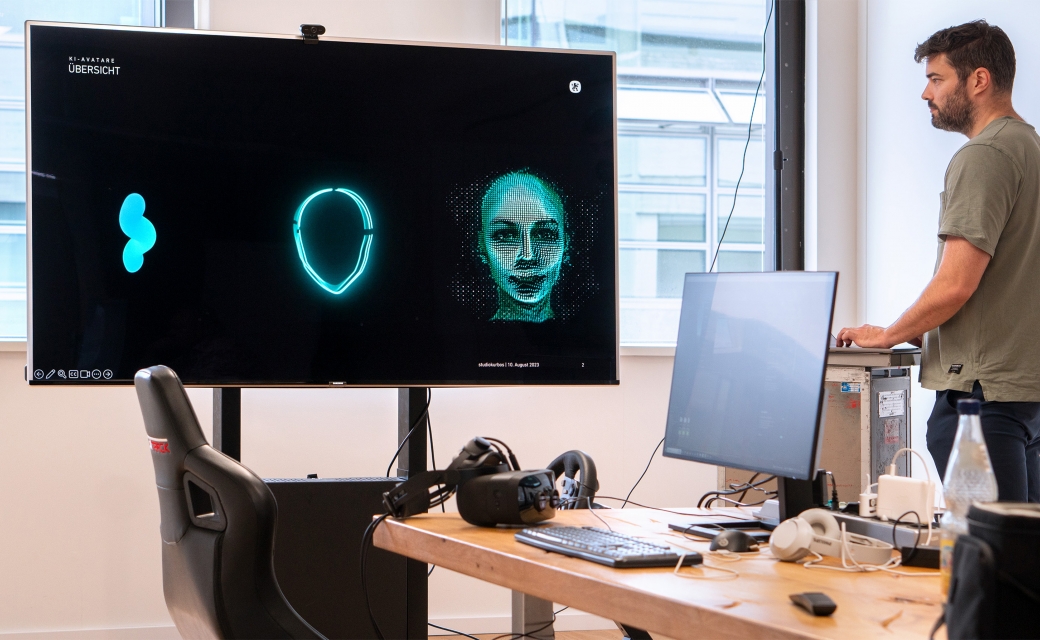 A-man-looks-at-a-large-screen-displaying-three-versions-of-mint-green-avatars-with-a-cable-car-seat-and-other-equipment-in-front-of-him